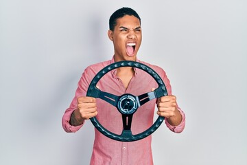 Young african american guy holding steering wheel angry and mad screaming frustrated and furious, shouting with anger. rage and aggressive concept.