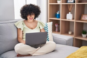 Poster - Young brunette woman with curly hair using laptop sitting on the sofa at home with a happy and cool smile on face. lucky person.