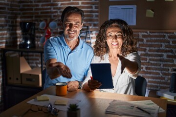 Canvas Print - Middle age hispanic couple using touchpad sitting on the table at night smiling cheerful offering palm hand giving assistance and acceptance.