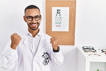 Canvas Print - African american optician man standing by eyesight test very happy and excited doing winner gesture with arms raised, smiling and screaming for success. celebration concept.