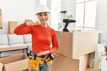 Poster - Middle age grey-haired woman wearing hardhat standing at new home gesturing with hands showing big and large size sign, measure symbol. smiling looking at the camera. measuring concept.