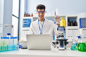 Sticker - Young hispanic man working at scientist laboratory doing video call smiling with an idea or question pointing finger with happy face, number one