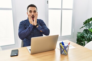 Poster - Young hispanic man with beard working at the office with laptop afraid and shocked, surprise and amazed expression with hands on face