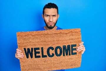 Hispanic man with beard holding welcome doormat skeptic and nervous, frowning upset because of problem. negative person.