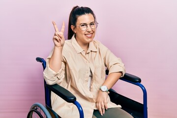 Poster - Young hispanic woman sitting on wheelchair smiling looking to the camera showing fingers doing victory sign. number two.