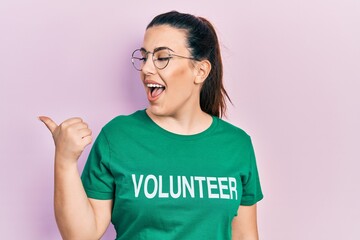 Sticker - Young hispanic woman wearing volunteer t shirt smiling with happy face looking and pointing to the side with thumb up.