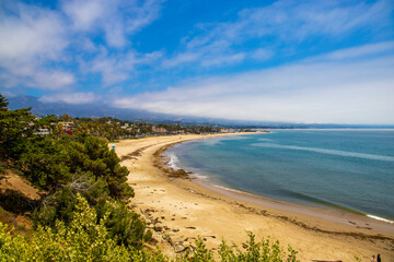 a gorgeous summer landscape at the beach with vast blue ocean water and silky brown sand surrounded by lush green trees, plants and homes with blue sky and clouds at Leadbetter Beach in Santa Barbara