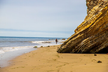 a man walking along the beach near the brown rocky cliffs with silky brown sand and blue ocean water with waves rolling into the beach with cloudy blue skies at Leadbetter Beach in Santa Barbara
