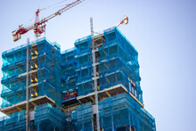 Two Multistory Buildings Facade Covered With Protective Construction Mesh On Blue Sky Background. Construction Of Residential Buildings, House Renovation, Skyscrapers In Big City. Construction Cranes.