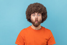 Portrait Of Silly Man With Afro Hairstyle In Orange T-shirt Puckers Lips Makes Grimace, Imitates Fish, Being Childish, Doing Funny Facial Expressions. Indoor Studio Shot Isolated On Blue Background.