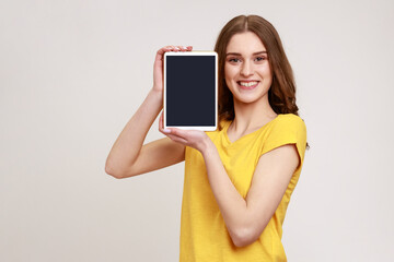 Wall Mural - Portrait of beautiful smiling young teenage with brown hair standing and holding tablet empty screen, sowing device and looking at camera. Indoor studio shot isolated on gray background.