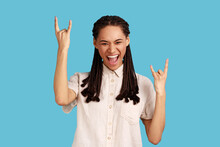 Happy Crazy Woman With Dreadlocks Showing Rock And Roll Gesture, Heavy Metal Sign, Enjoys Favorite Music On Party, Has Squints Face, Exclaims From Joy. Indoor Studio Shot Isolated On Blue Background.