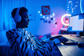 Arican teenage boy in wireless headphones playing video game on computer while sitting at table in dark room