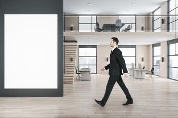 Poster - Side view of businessman walking in modern two floors wooden and concrete duplex office interior with empty white mock up poster on wall, panoramic city view, furniture and daylight.