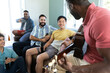 © wavebreak3 - Multiracial happy male friends with beer bottles and cellphone looking at man playing guitar at home
