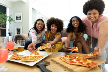 excited biracial female friends picking pizza slices on table while spending leisure time at home