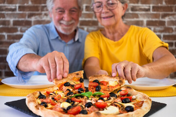 Wall Mural - Defocused senior couple sitting at home table taking a slice of pizza enjoying delicious food