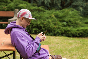 young caucasian man wears a violet sweatshirt and cap sitting on park bench and using smartphone. pe