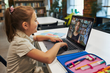 Student litttle girl having video class remotely with her teacher on laptop sitting at desk in after school club at primary school. Back to school. Child using technology