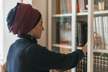 Religious Jewish Young Woman With Head Covered Stands Near Bookcase With Religious Books (9)
