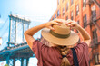© LiettaOraz - Fashionable tourist woman visiting New York City wearing hat and stylish street style outfit. Young girl amazed by the beautiful city. Manhattan bridge view on summertime.