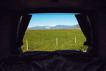 Looking out the camper minivan back from built in bed. Traveling Iceland with van. View over mountains, pasture and glacier, blue sky, sunny day.