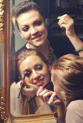 Two smiling young women putting make up in front of the mirror before going out