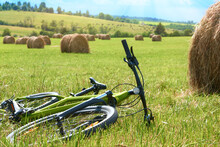 The Bicycle Lies On The Grass In A Field Near A Stack, Overlooking A Pasture. Summer Cross-country Bike Ride. Rest After A Bike Ride.