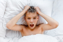 Worried Awake Teen Boy In A Bed Is Shocked Because It Is Too Late. Kid Lies On The Pillow And Clutched His Head In Horror Because He Is Overslept