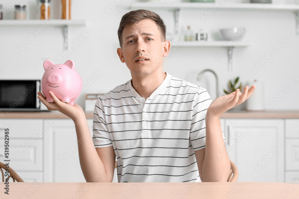 Upset young man with piggy bank in kitchen