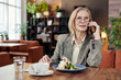 Mature businesswoman in eyeglasses talking to her clients on mobile phone while sitting at the table and eating dessert at the restaurant