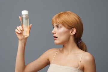 Horizontal studio shot. A cute woman in a white T-shirt with light clean skin, with red silky hair gathered in a ponytail on a greyish background with a transparent bottle of water.