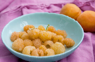 Yellow raspberries in a turquoise bowl on a pink fabric near a pair of apricots