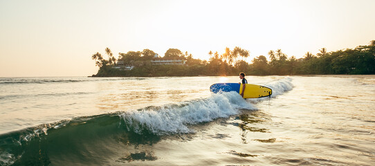 Wall Mural - Young teenager boy with colorful surfboard go to foamy sea for surfing. He enjoying a beautiful sunset light on Dewata beach on Sri Lanka. Active sport vacation and exotic countries traveling concept.