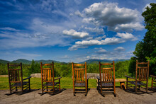 Row Of Wooden Rocking Chairs Looking Out Over A Green Landscape And Blue Sky With Puffy Clouds.
