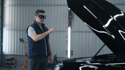 A male mechanic in a virtual reality headset gesturing and standing near the open hood of a car, while making a diagnostics in a car service