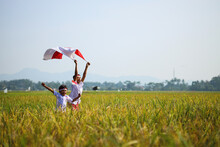 Indonesian School Students Wearing Uniform Are Raising Their Hands While Holding Red White Flag In The Midst Of The Rice Field. Celebrating Independence Day.