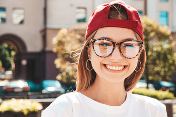 Young beautiful smiling hipster woman in trendy summer clothes. Sexy carefree woman posing on the street background in cap at sunset. Positive model outdoors. Cheerful and happy in spectacles