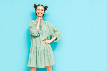 Young beautiful smiling female in trendy summer green dress. Sexy carefree woman with two horns hairstyle posing near blue wall in studio. Positive model having fun. Cheerful and happy