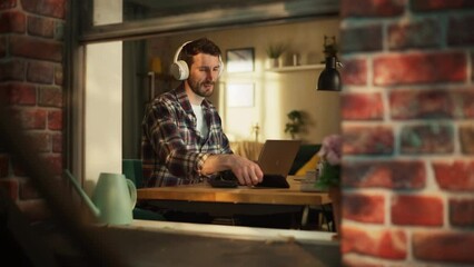 Aufkleber - Handsome Caucasian Man Working on Laptop Computer while Sitting Behind His Desk at Home Office. Freelance Entrepreneur Listening to Music, Doing Remote Telework. View From Outdoors into Window.