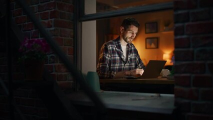 Aufkleber - Caucasian Man Using Laptop While Working Remotely at Home Office. Handsome Male Sitting at Table, Works On Computer, Brainstorms Creative Project, Browsing Internet. View From Outdoors into Window.