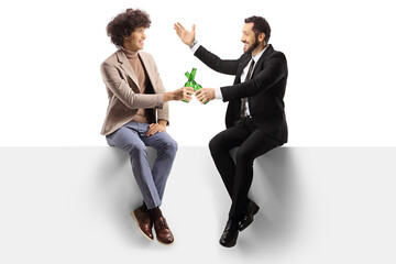 Wall Mural - Cheerful young men sitting on a blank panel and toasting with bottles of beer