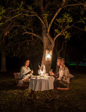 Couple On Safari In South Africa, Asian Women And European Men Having A Bush Dinner With Candlelight At Night. Romantic Dinner At Night In The Bush During A Luxury Safari In South Africa