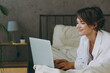 Young happy woman wear white shirt pajama she lying in bed hold use work on laptop pc computer study online browsing internet rest relax spend time in bedroom lounge home in own room hotel wake up