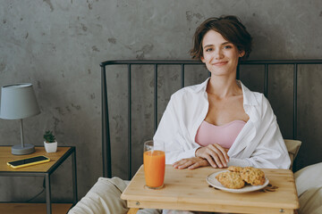 Wall Mural - Young smiling happy woman wear white shirt pajama she lying in bed eat breakfast look camera rest relax spend time in bedroom lounge home in hotel room wake up dream be lost in reverie good mood day.