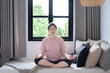 © Bond_JP - Cheerful young Asian woman in pink t-shirt and leggings sitting in Ardha Padmasana position on couch near windows, practicing yoga, doing half Lotus pose with mudra gesture. Yoga,well-being concept.