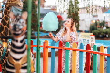 Wall Mural - Summer festival. Dreamy pretty blonde woman holding a cotton candy at amusement park. The concept of vacation, holiday and happiness