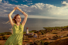 A Young Carefree Girl Stands On A Mountain With Her Hands Raised Up At Sunset, A Lighthouse Is Visible Below