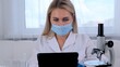 © Евгения Медведева - female research chemist scientist in a protective mask and gloves, dressed in a medical suit, with a tablet computer and a microscope, conducts research in the laboratory.