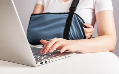 Poster - Woman with painful arm in sling working on laptop. Female wearing bandage to support injured elbow, shoulder, forearm or wrist while sitting at table with computer. High quality photo
